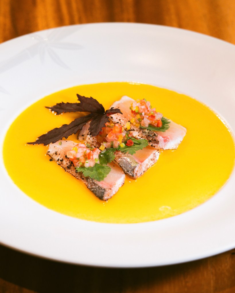 A beautifully plated dish featuring slices of fish, topped with colorful diced vegetables and herbs, served in a vibrant orange sauce.
