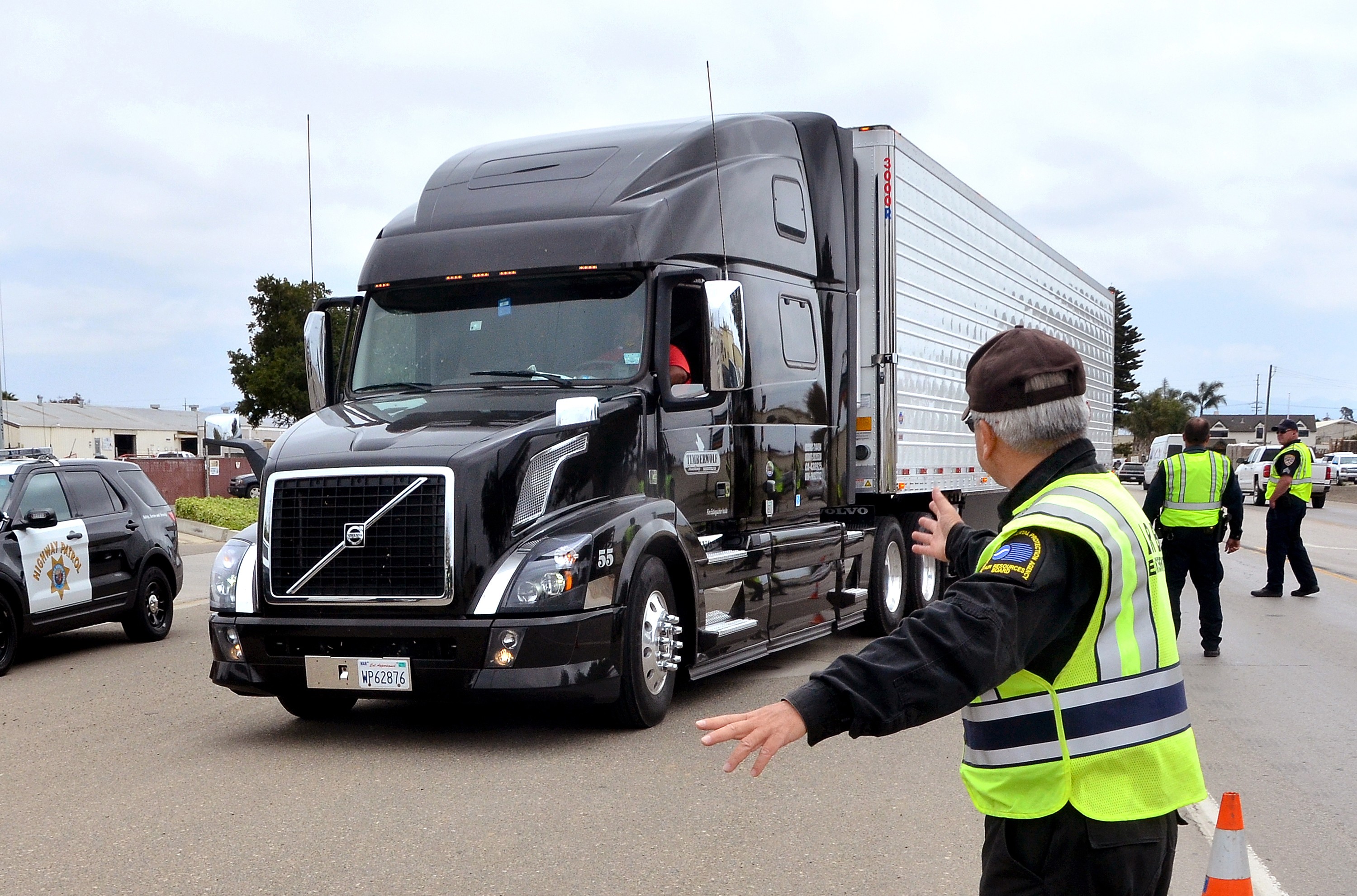 An enforcement officer wearing a high-visibility yellow safety vest directs a large black Volvo semi-truck at a roadside inspection checkpoint. A California Highway Patrol vehicle is visible on the left, and additional officers in safety vests stand in the background. Traffic cones mark the checkpoint area.