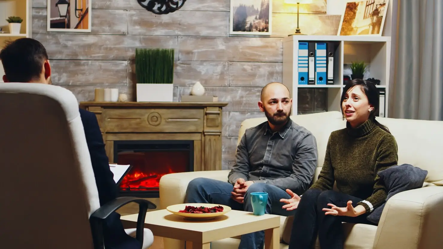 Couple in a therapy session with a male counselor in a comfortable room with a fireplace.