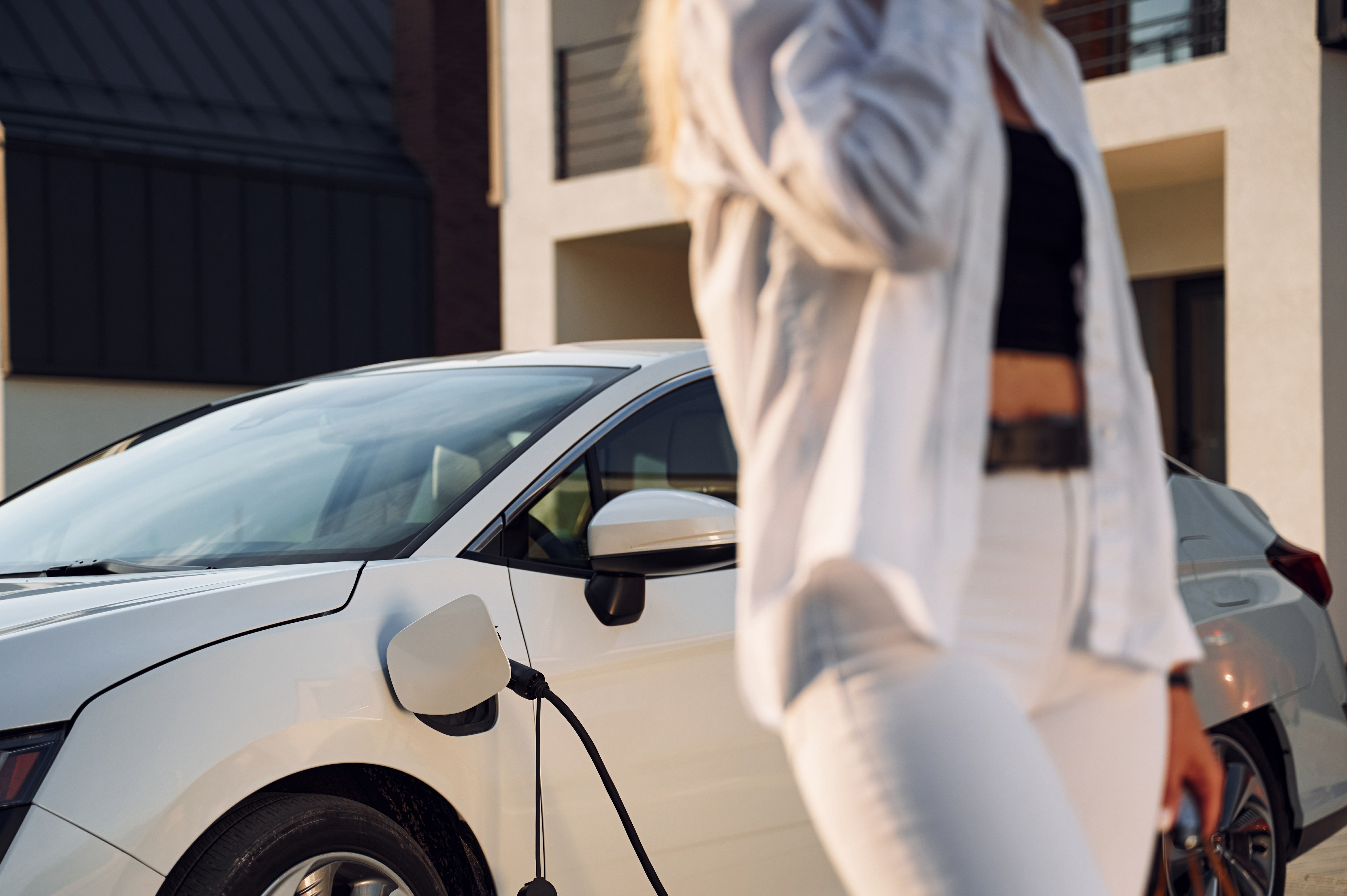 Young woman in white clothes is with her electric car with a shopping bag at daytime.