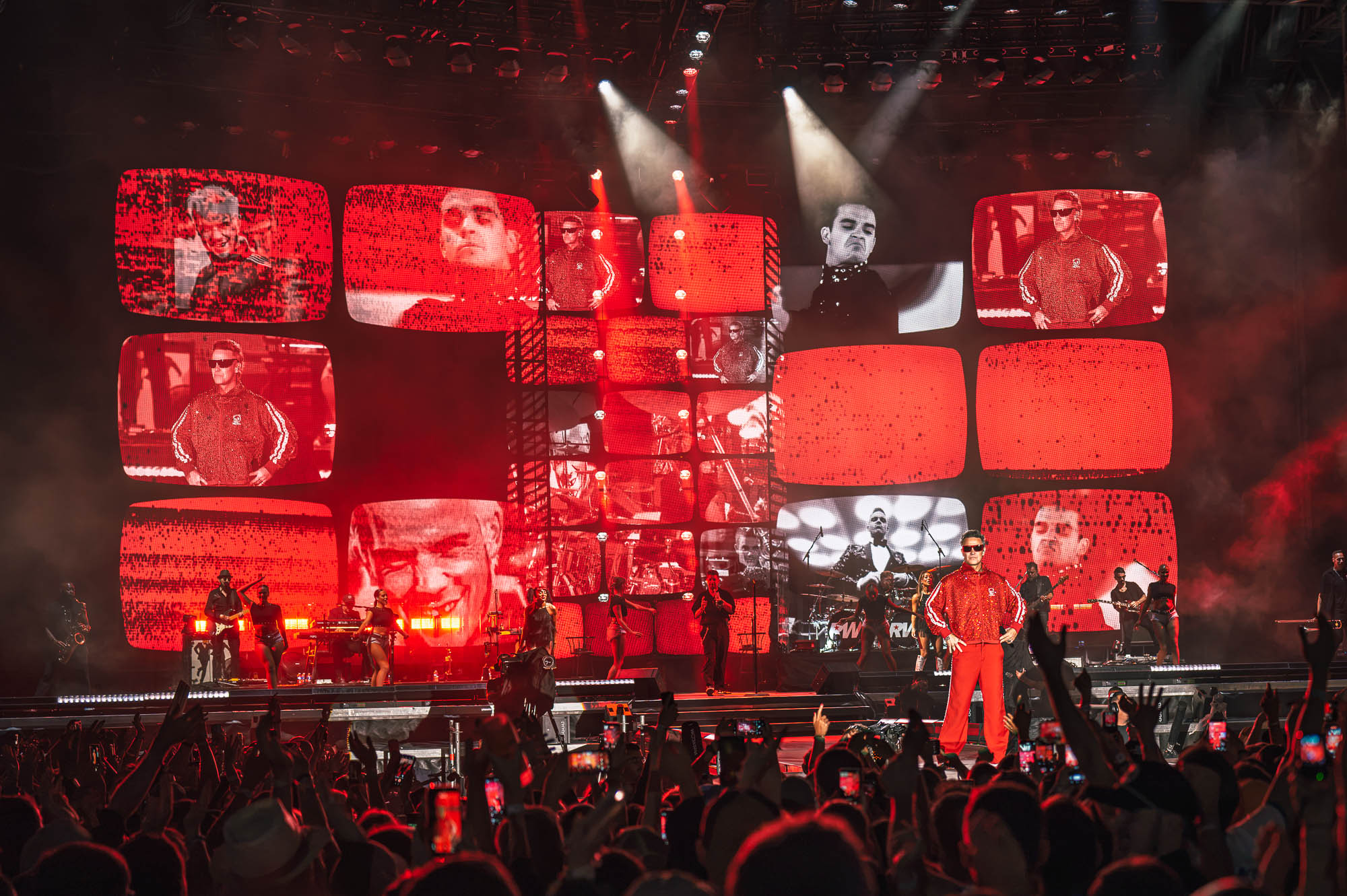 Robbie Williams en tenue rouge face au public, entouré par un immense mur d’écrans façon téléviseurs, sous une lumière rouge.