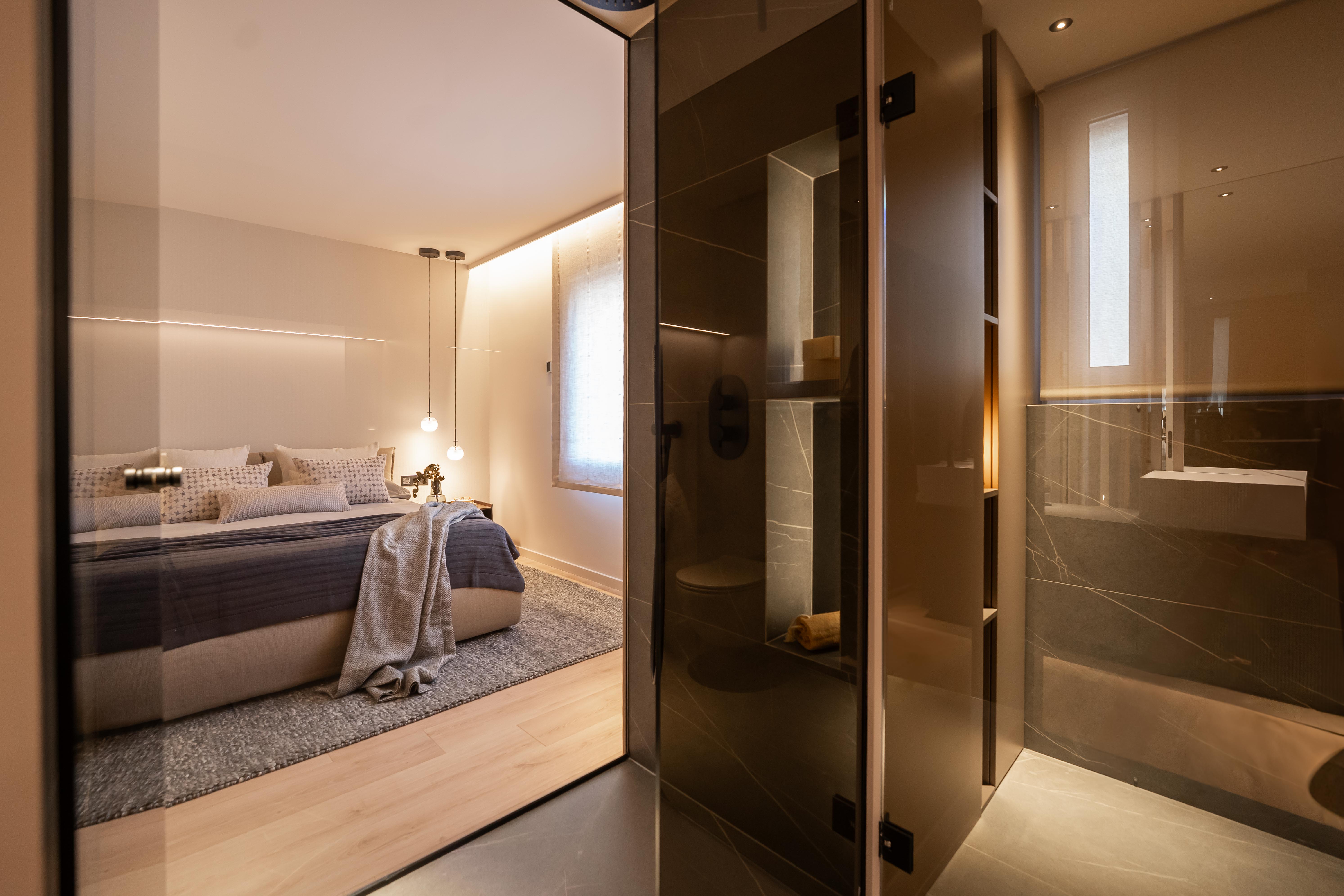 Dormitorio minimalista con tonos grises visto desde cristal del baño en suite