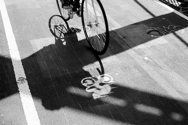 A black and white photograph captures a bicycle in motion, viewed from below. The bicycle's front wheel is prominently featured, with its shadow cast across the asphalt. The asphalt surface has white painted lines, and a dark circular marking is visible near the right edge of the frame. The photograph is taken from a low angle, emphasizing the bicycle's height and providing a dynamic perspective of the bike in motion.