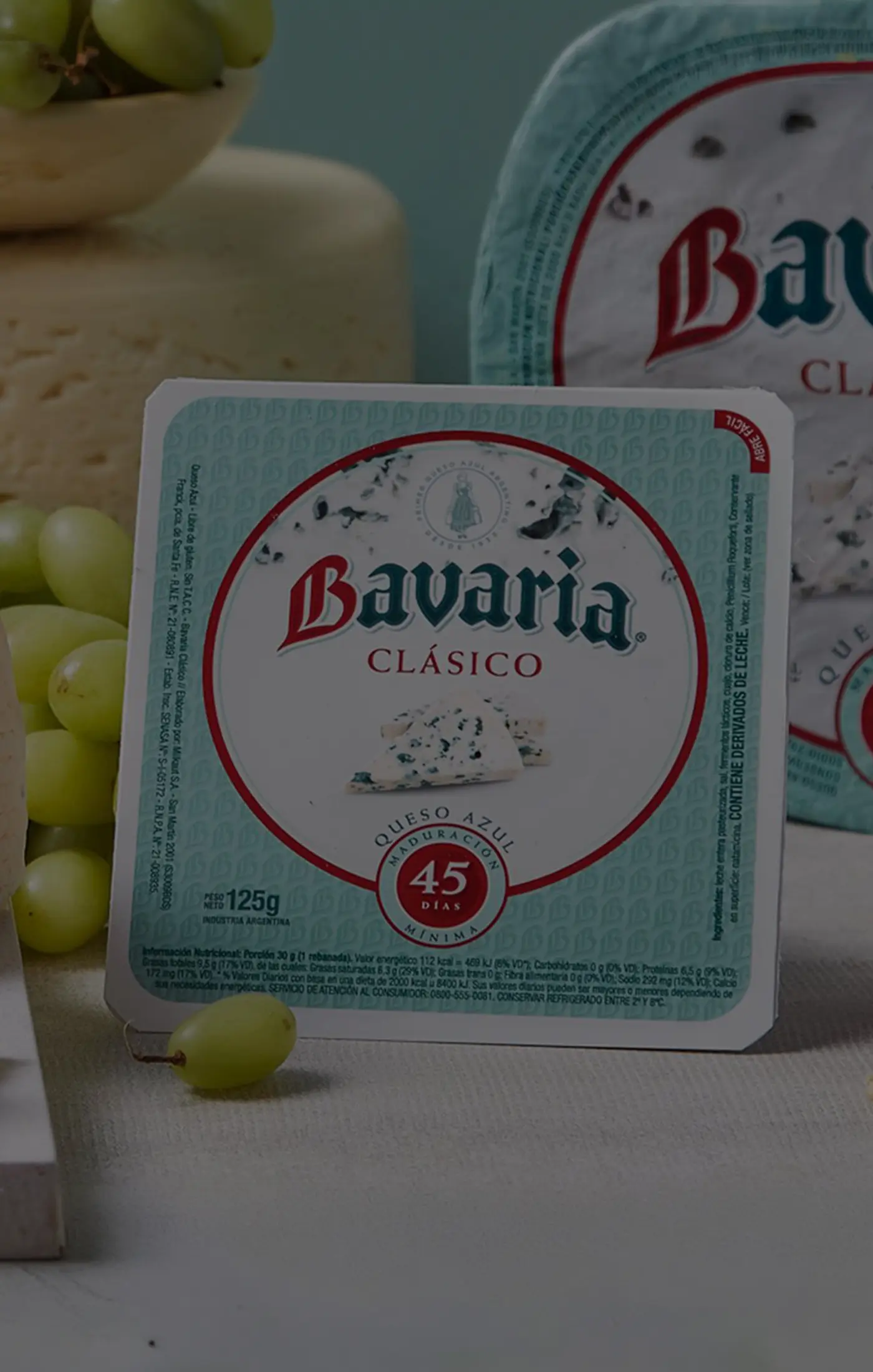 Packaging and slices of Bavaria Clásico blue cheese are surrounded by fresh green grapes and a small pile of pistachios on a table. The scene conveys a gourmet and inviting atmosphere, highlighting the cheese's 45-year tradition.