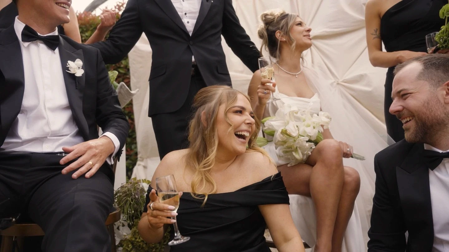 A joyful group of elegantly dressed people celebrate at a wedding, with one person holding a bouquet of white flowers and others clinking glasses of champagne, set against a backdrop of draped white fabric.