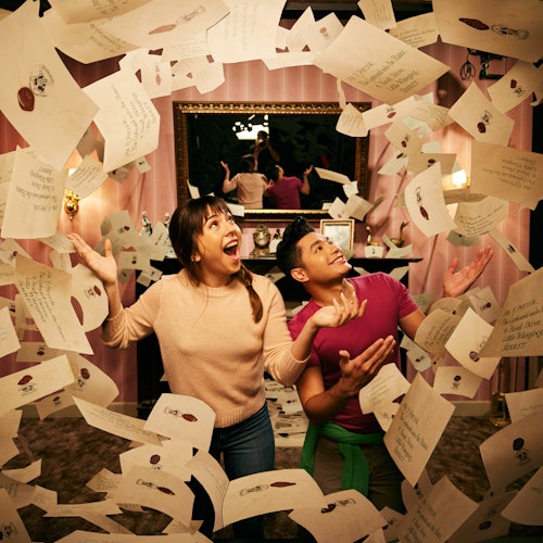 Two people joyfully surrounded by flying envelopes in a decorated room with a mirror and pink wallpaper.