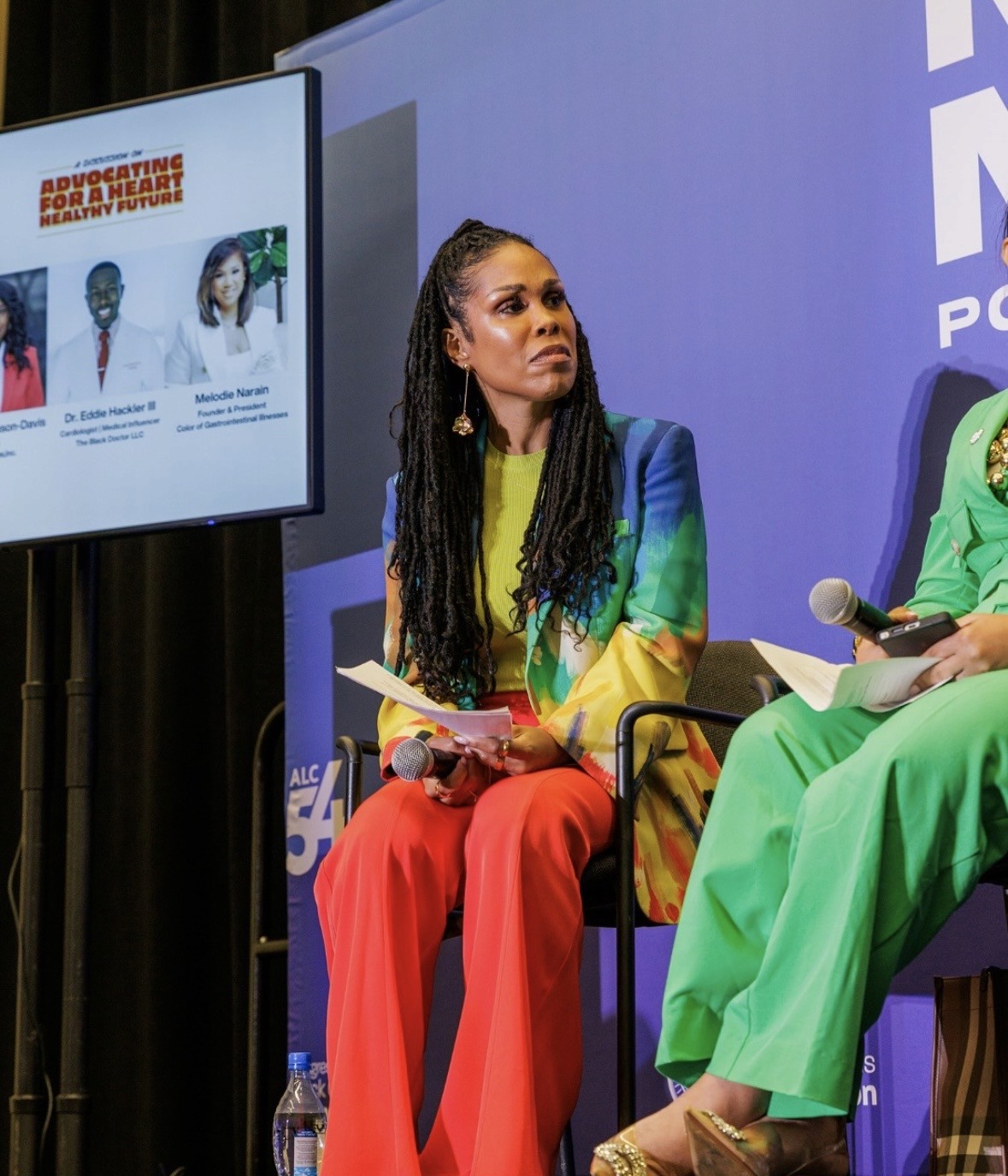 A woman in a colorful suit sits onstage holding papers, attentively listening during a panel discussion. A screen beside her displays speaker names and photos under the heading Advocating For A Healthier Heart + Healthier Future.