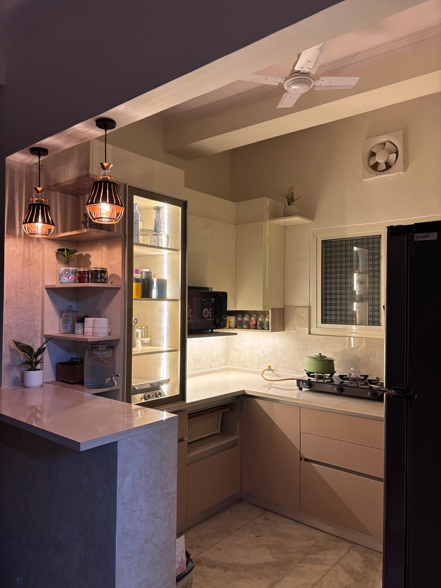 Blush-toned modular kitchen with pendant lights over a breakfast counter and open corner shelving