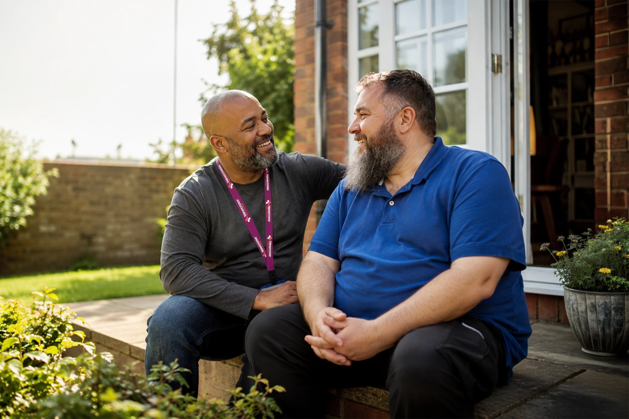 Mental health support worker sharing a warm, supportive moment with a bearded man as they smile together in a sunny residential garden.