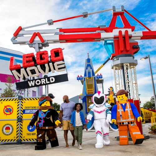 A family poses with Lego characters under a "Lego Movie World" sign in a theme park setting.