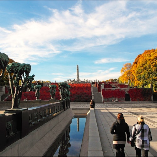 People walking along a pathway lined with sculptures and trees with autumn foliage, under a sunny blue sky.