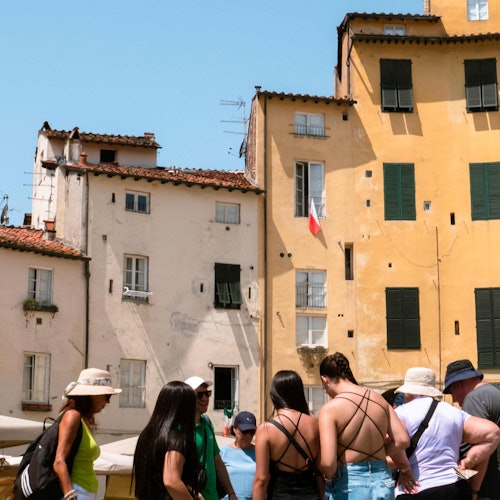 Un gruppo di persone in abiti estivi si raduna davanti a edifici storici con facciate gialle e bianche sotto un cielo blu limpido.