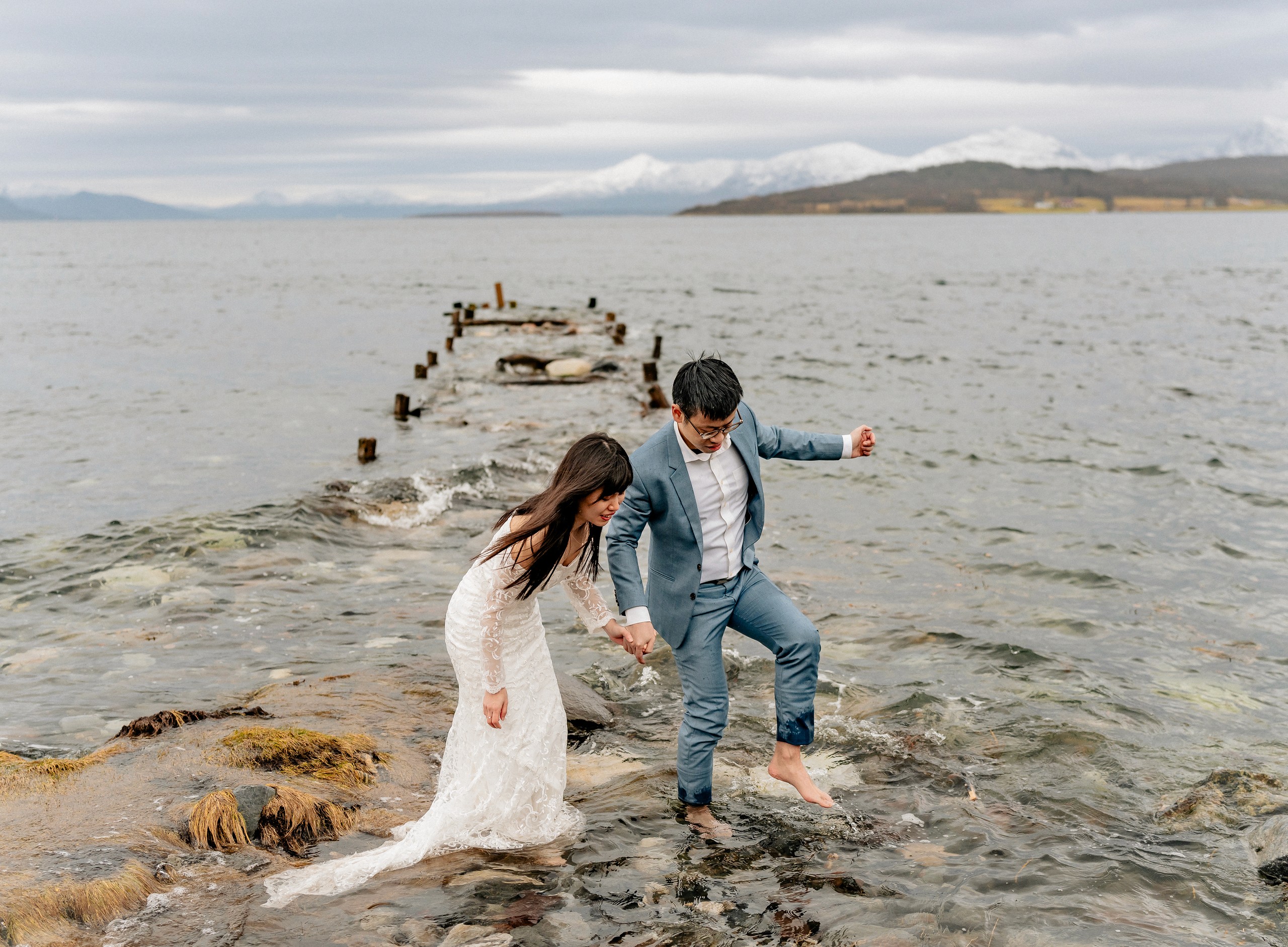 A couple stepping barefoot across exposed rocks and shallow water beside a weathered pier.