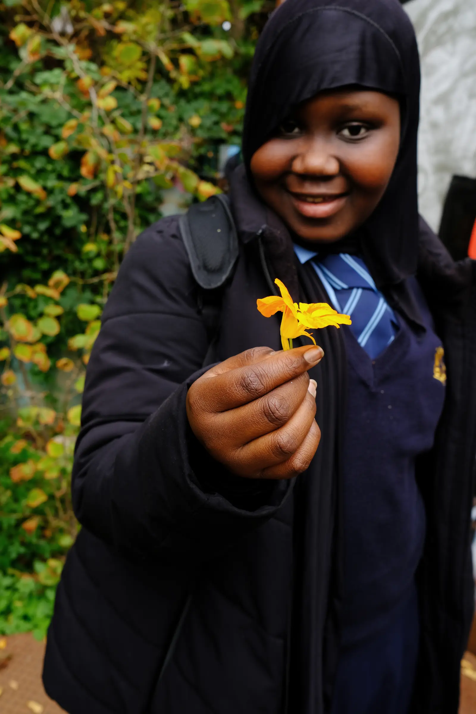 A person smiling broadly while holding a yellow object, surrounded by greenery in the background.