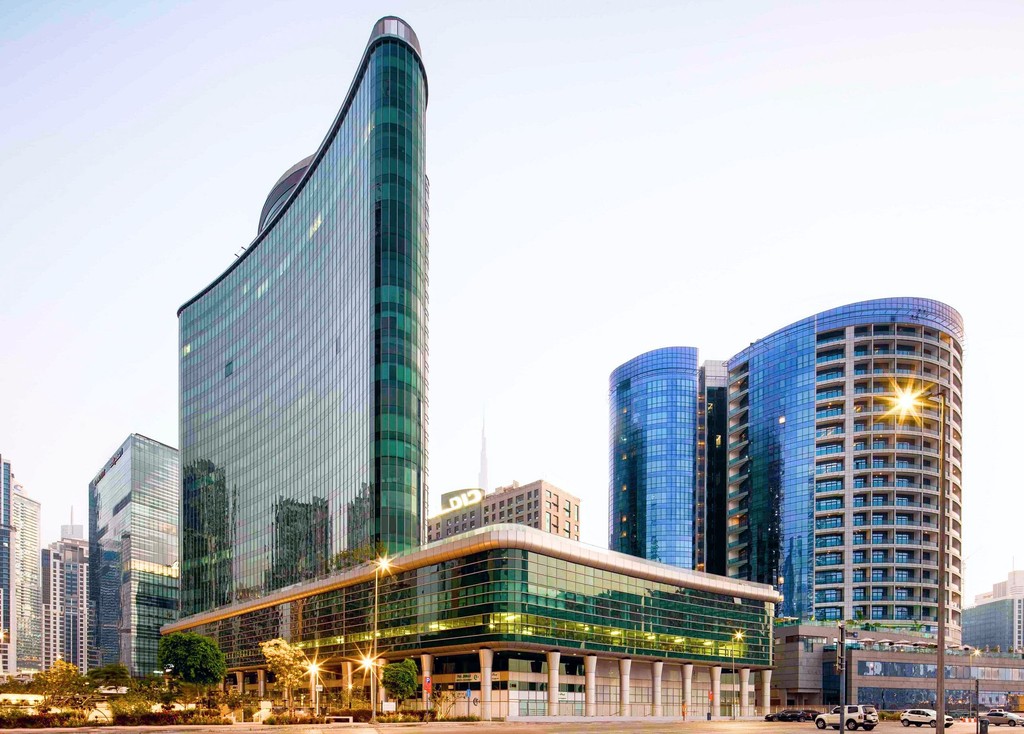 A large building with a green glass facade, known as The Binary by Omniyat, situated in a bustling city center.