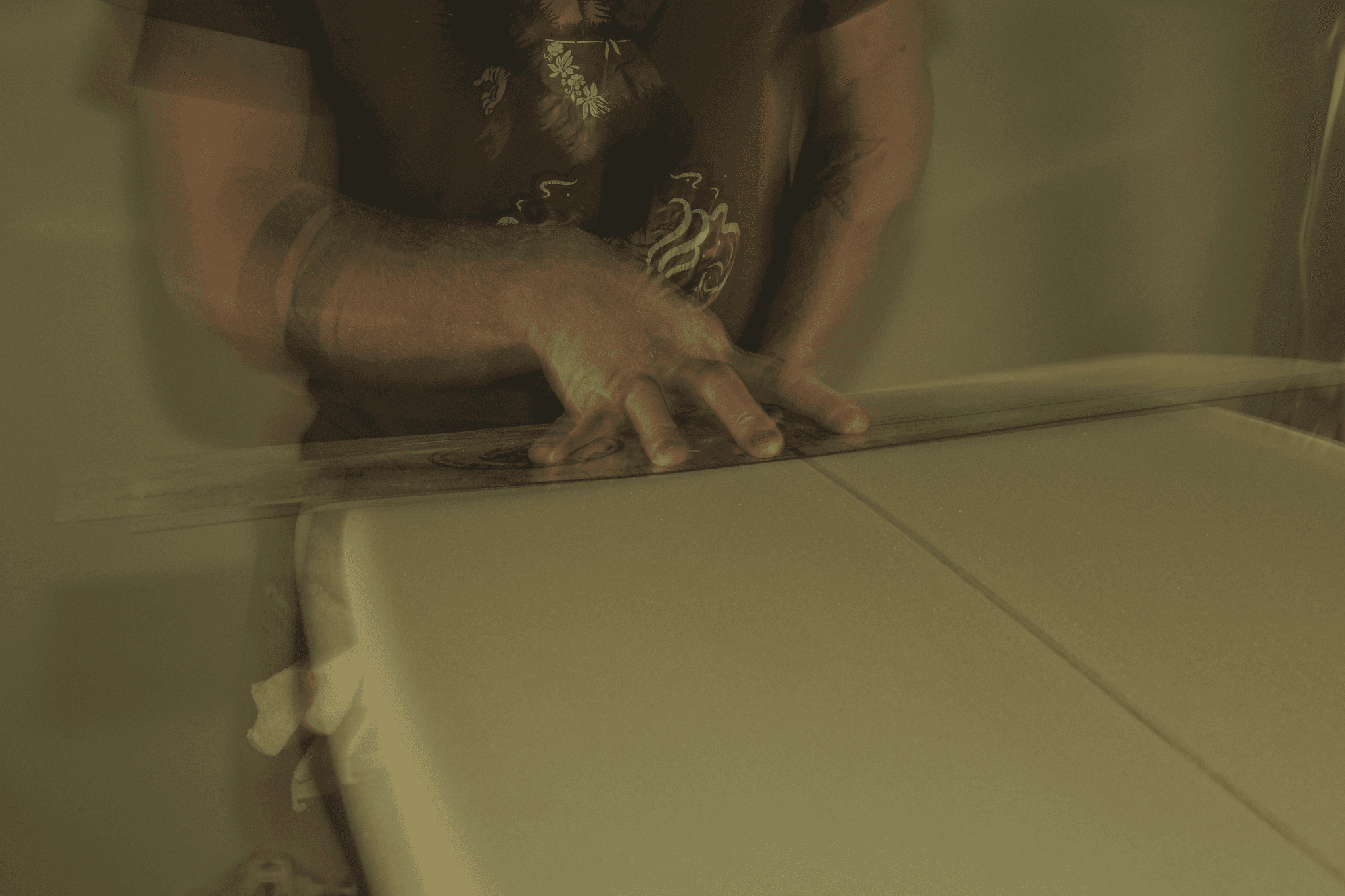 Primo piano sulle mani di uno shaper che utilizza un righello trasparente per misurare o tracciare una linea su un blank di schiuma per la tavola da surf.
