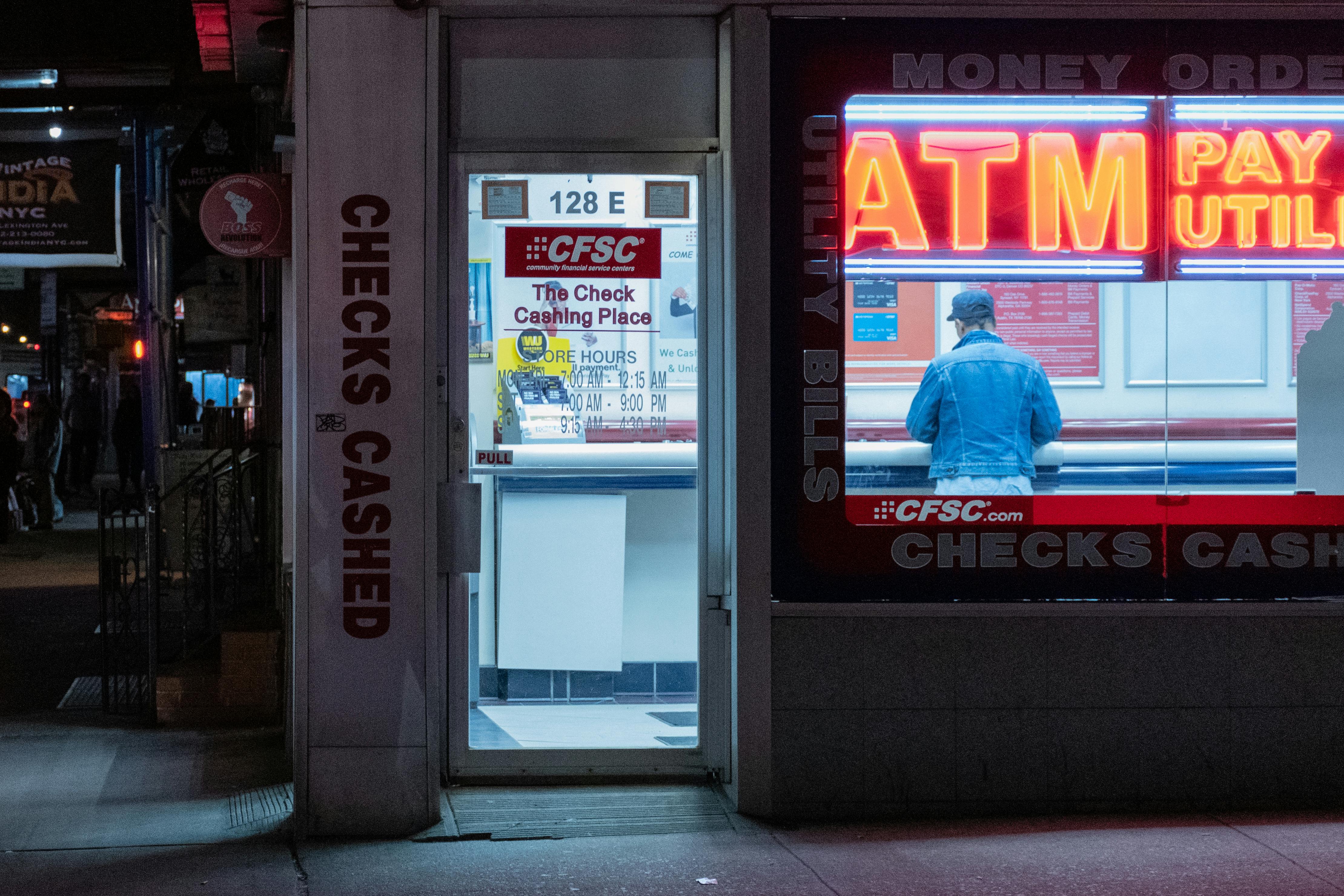 A person is standing at an ATM in a dimly lit street, next to a bright CFSC storefront that advertises services like check cashing and utility bill payments, illuminated by neon signs.