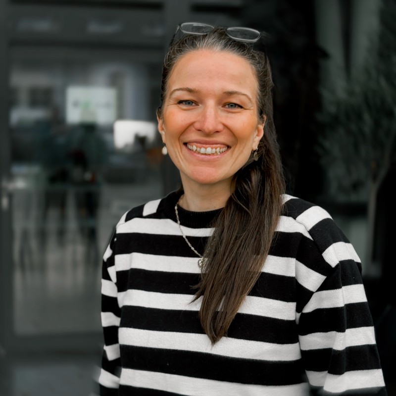 A young woman with long hair smiles while standing against a green backdrop, wearing a casual dark sweater.