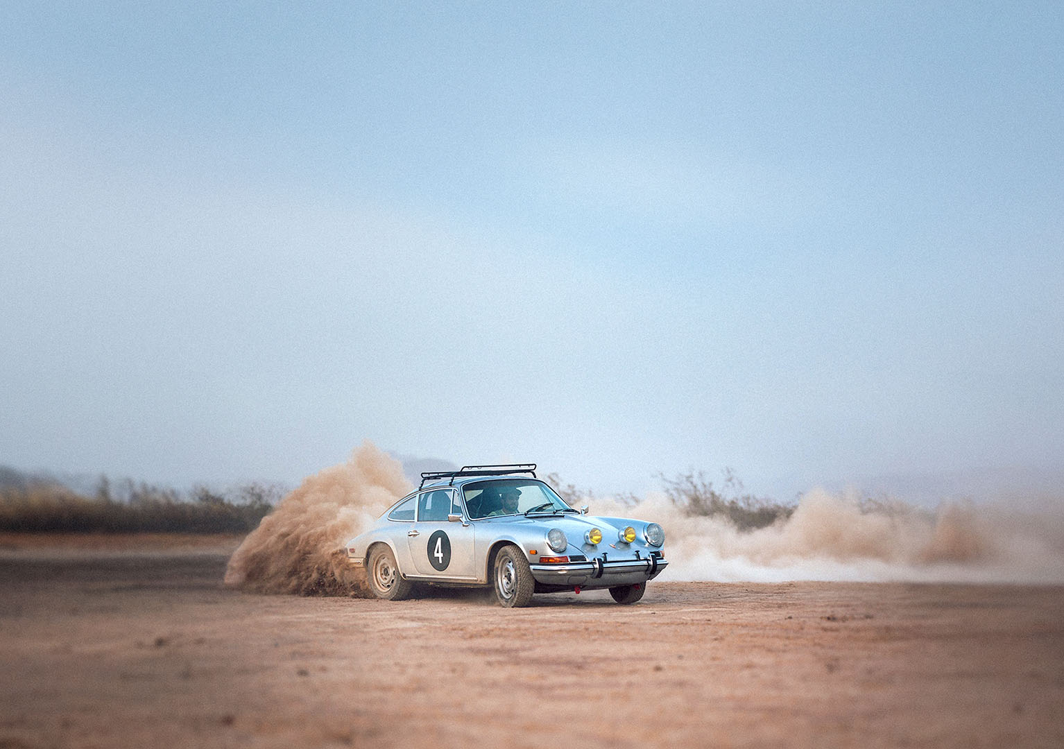 A vintage silver Porsche rally car with the number 4 on its side kicks up a cloud of dust as it drifts on a dirt track in a desert landscape.