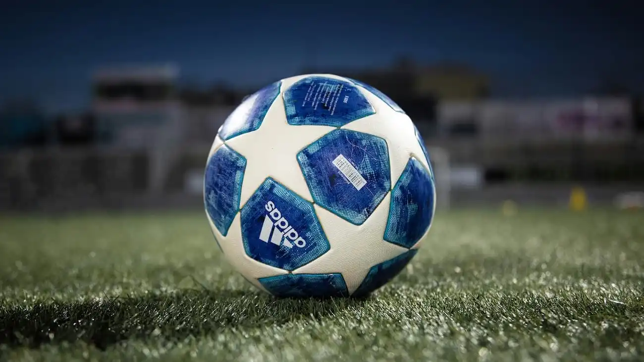 This image captures the official Adidas match ball of the UEFA Champions League, poised on the lush green turf under the stadium lights, symbolising the anticipation and excitement of the upcoming 2024/25 season. The ball, adorned with a distinctive blue star pattern, represents not only the pinnacle of football performance but also the dawn of a new era as the Champions League embraces significant changes to its format, promising a more dynamic and inclusive tournament for clubs across Europe.