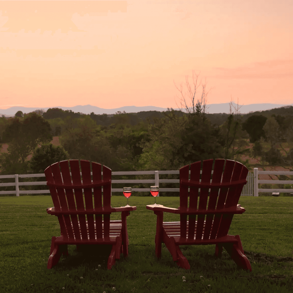 Two red Adirondack chairs face a scenic countryside view at sunset, with two glasses of pink wine resting on the armrests, embodying a serene outdoor setting ideal for a relaxing time on the patio or porch.