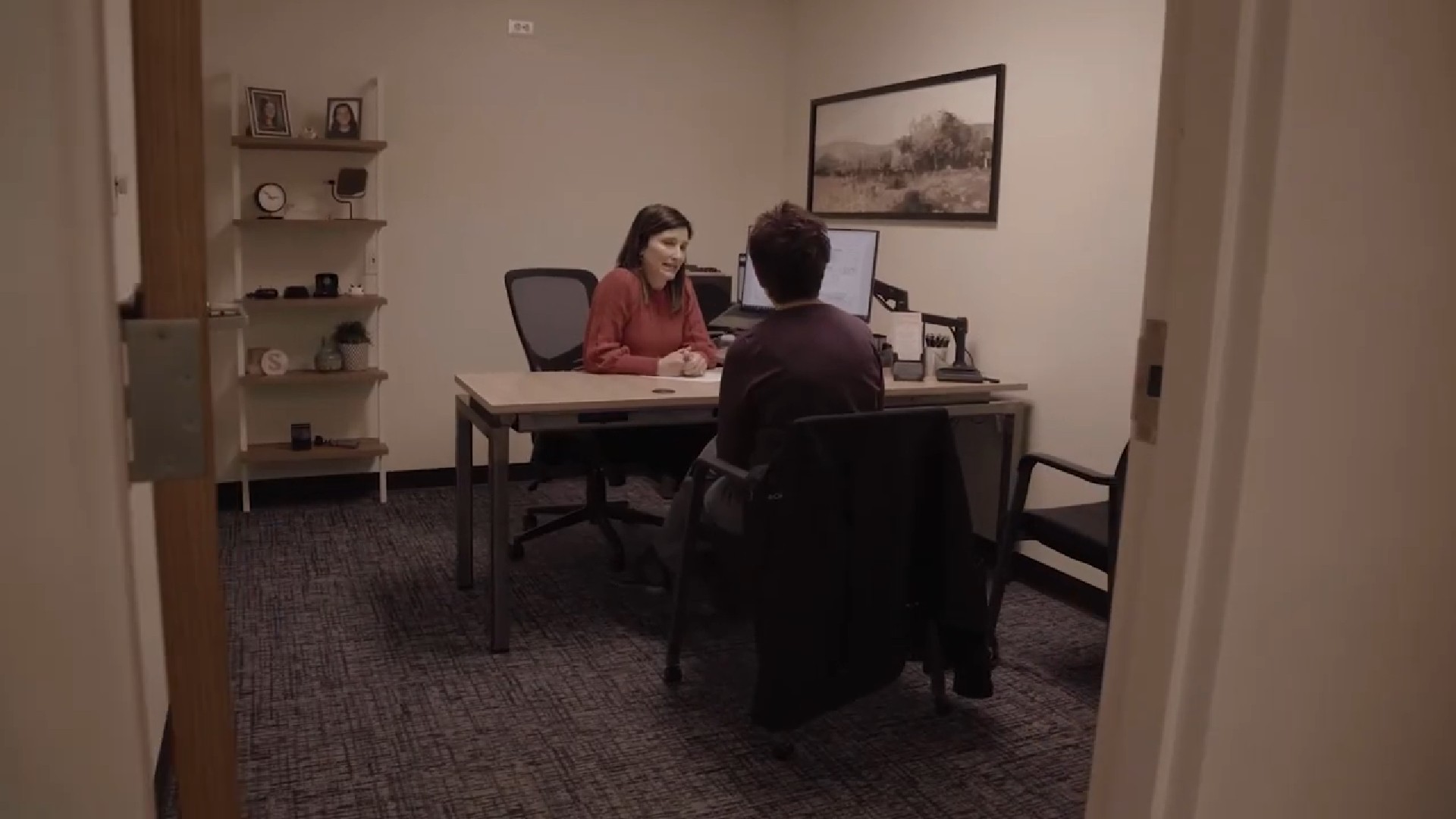 Two people work at desks in an office, with computers and shelves in the background.