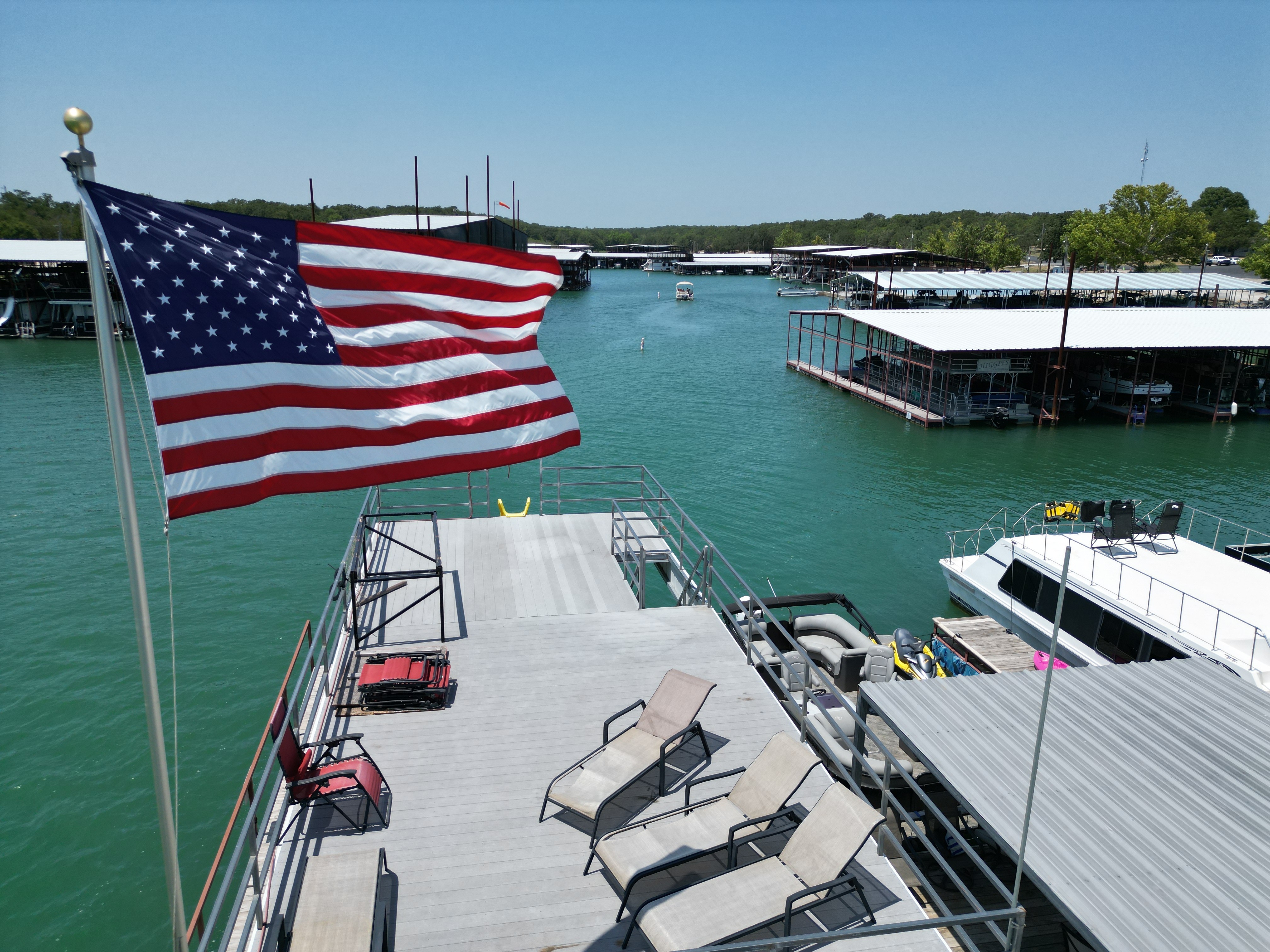 A vibrant American flag waves above a docked pontoon boat on a serene lake, surrounded by several metal-roofed boat docks and lush green trees in the background, under a clear blue sky.