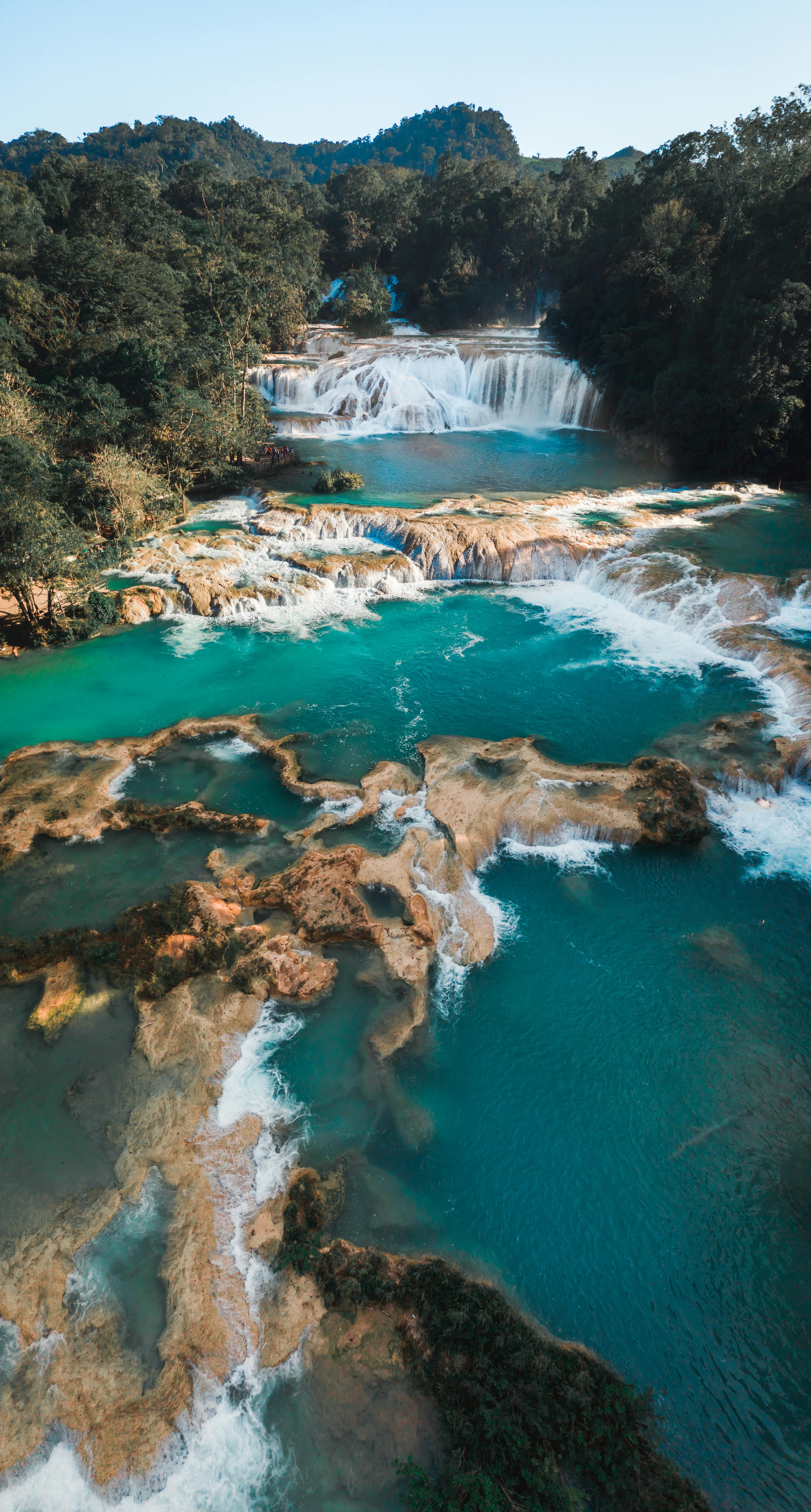 Aerial view of a cascading waterfall surrounded by lush green forest and turquoise waters, showcasing the natural beauty and tranquility of the tropical landscape.