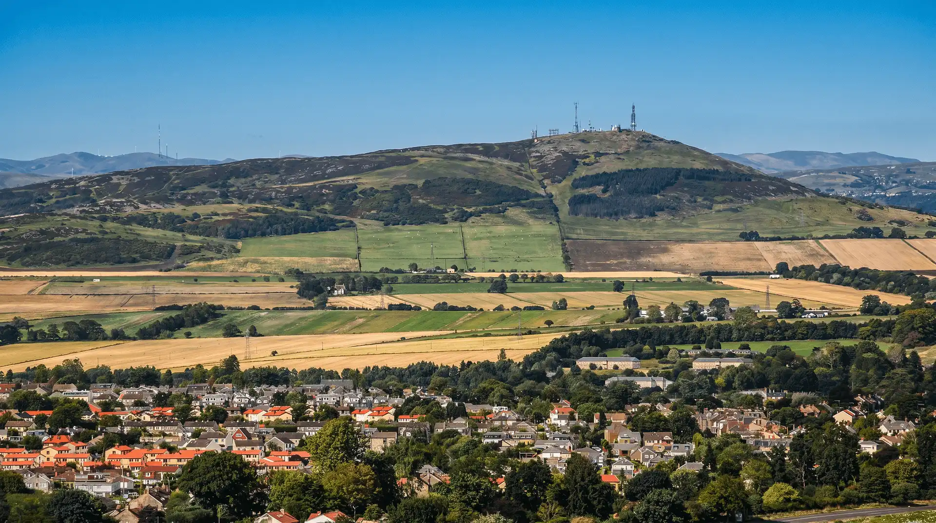 A layered panoramic view showing a residential town in the foreground with colorful rooftops and trees, followed by agricultural fields in shades of green and gold. In the background, a high hill features transmission towers beneath a clear blue sky.