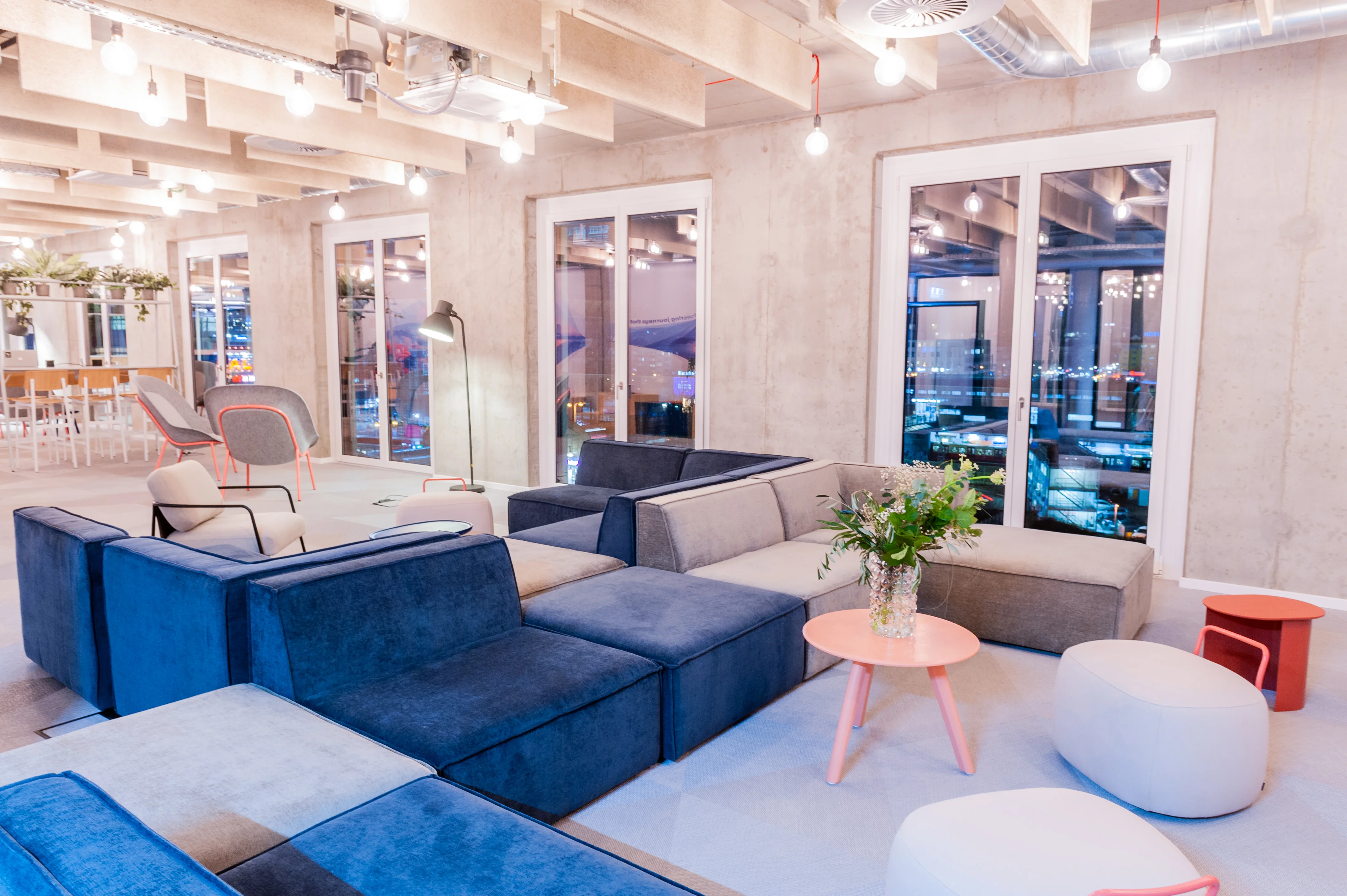 Modular lounge at OMIO with navy and light grey velvet sofa blocks arranged back to back, a peach round coffee table with flowers, poufs and tall French windows in a concrete facade.