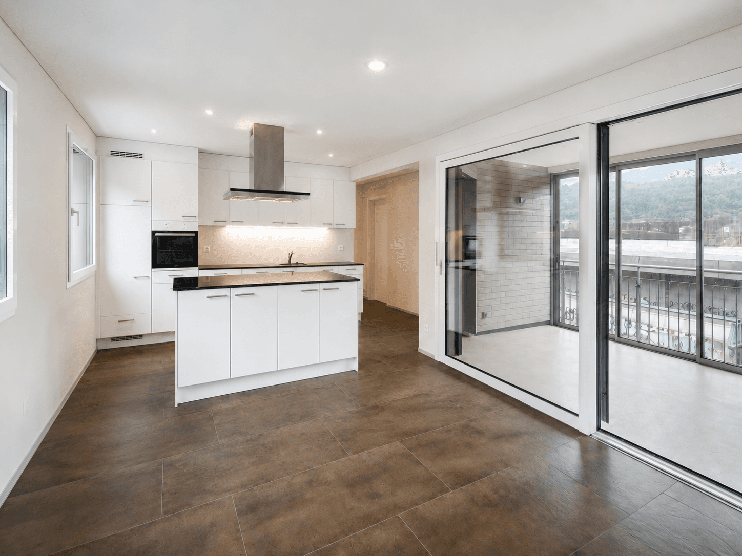 Heller, leerer Wohnraum mit offener weisser Küche, Kochinsel, dunklem Plattenboden und grosser Fensterfront zum gedeckten Balkon.