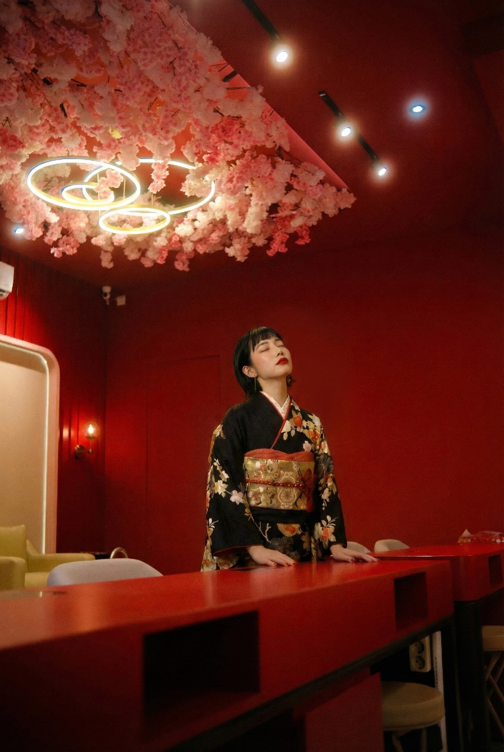 Woman in a kimono looks up under a ceiling of pink blossoms. A red nail bar and lights illuminate the scene.