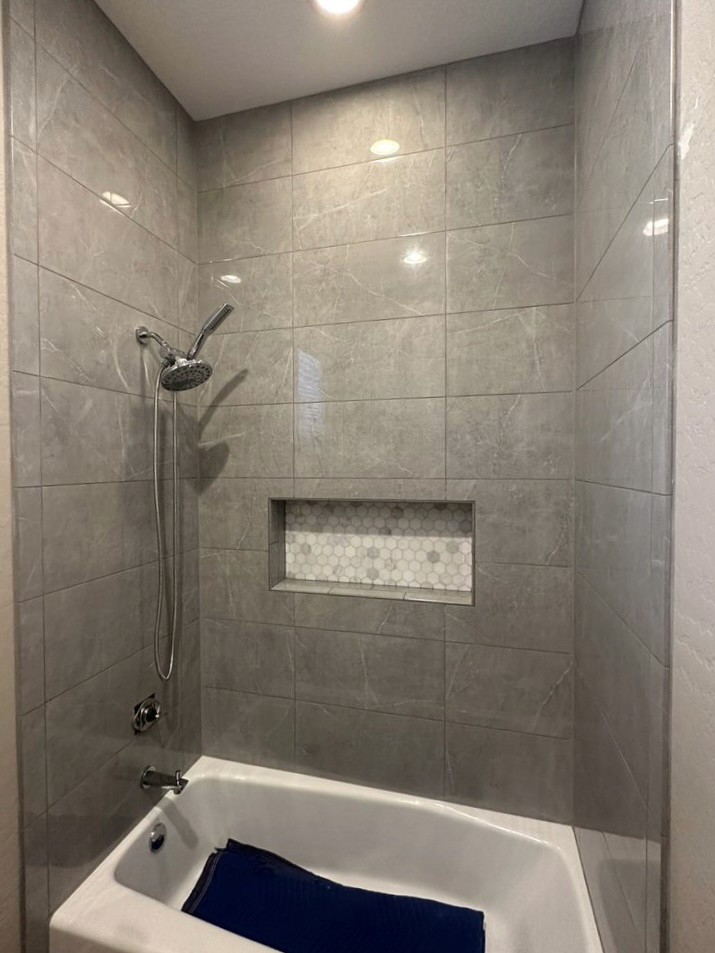 Bathtub shower combo with sleek grey marble-look large format wall tiles, a built-in shower niche with a white and grey hexagonal mosaic tile accent, and modern chrome fixtures.