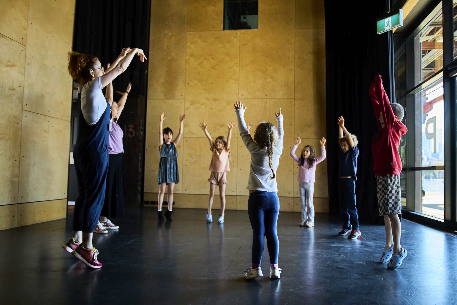 A group of young children stand in a circle with a teaching artist, all raising their arms during a movement warm-up in a studio.