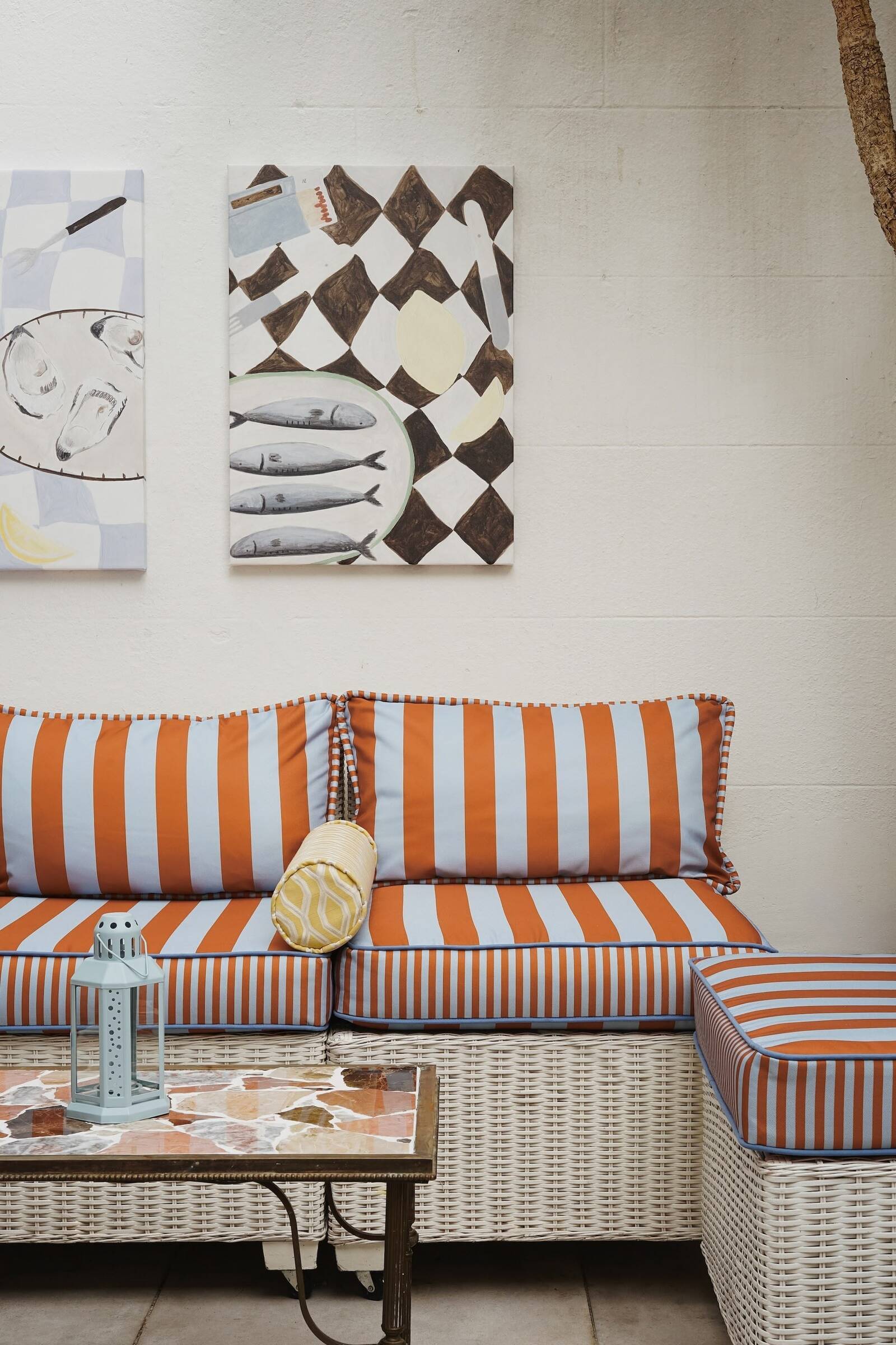 Beach-themed patio seating area with a wicker sofa with orange and white striped cushions, a small marble-topped table with a lantern, and wall art depicting fish and seafood, proving that outdoor furniture can be elevated.