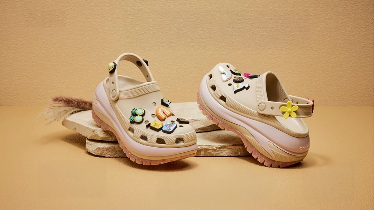 Crocs Classic Mega Crush clogs in cream and pink displayed on stone slabs, decorated with colourful Jibbitz charms including rainbow, flower and tropical designs, warm beige studio background
