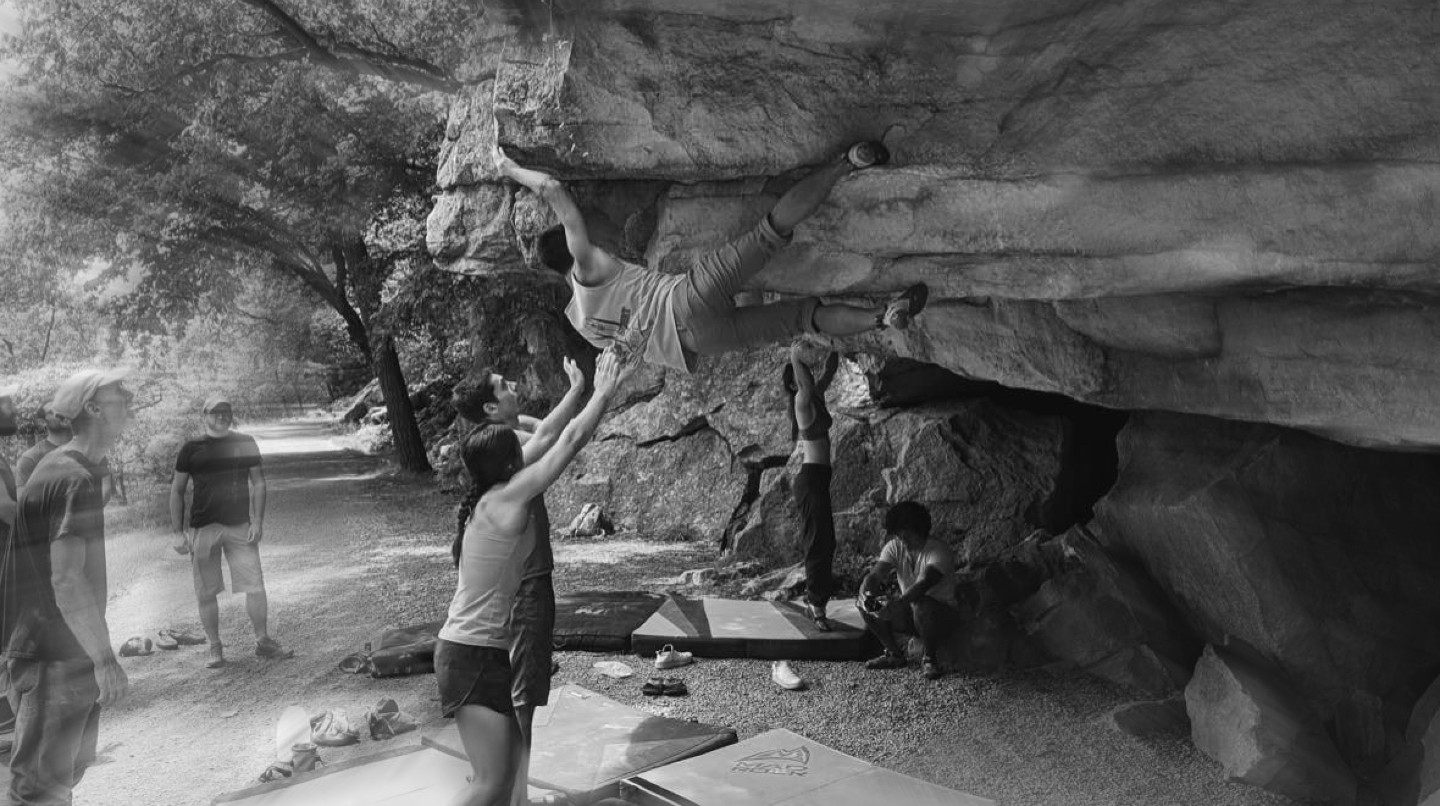 Black and white photo of a rookie rock climber in an uncomfortable position supported from the ground by the hands of friends 