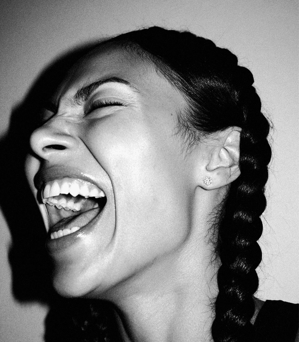 Black and white close-up portrait of a laughing woman with braided hair, dramatic shadows, and expressive editorial photography style