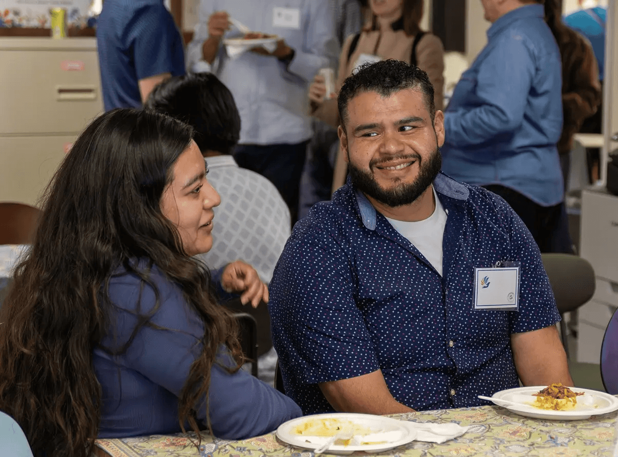 A man and a woman are seated at a table in a social gathering, engaging in conversation with smiles, as other attendees interact in the background, creating a warm and inviting atmosphere.