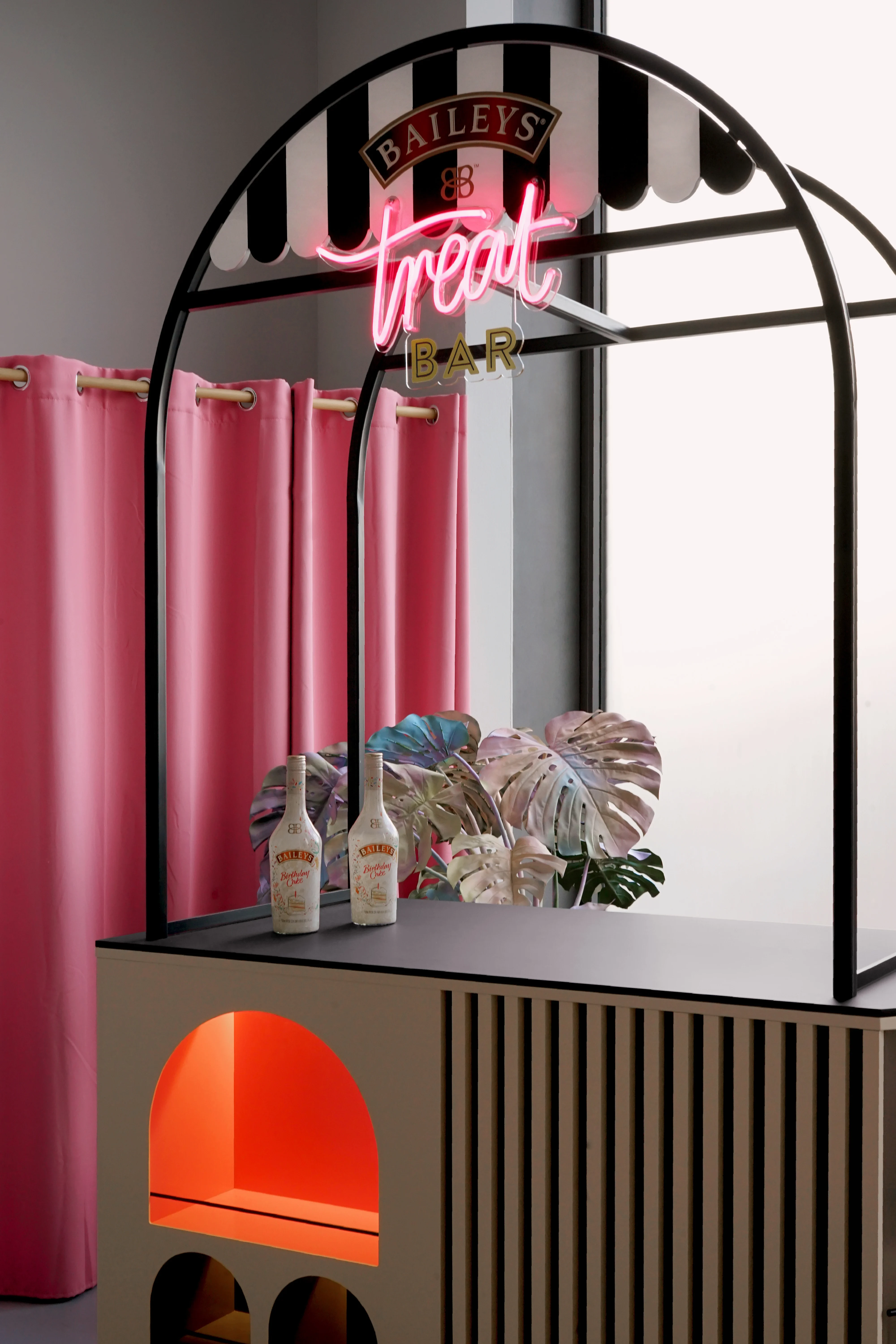 Black arch frame of the BAILEYS Treatbar with striped awning, pink treat neon sign and Baileys bottles on the counter next to iridescent monstera leaves.