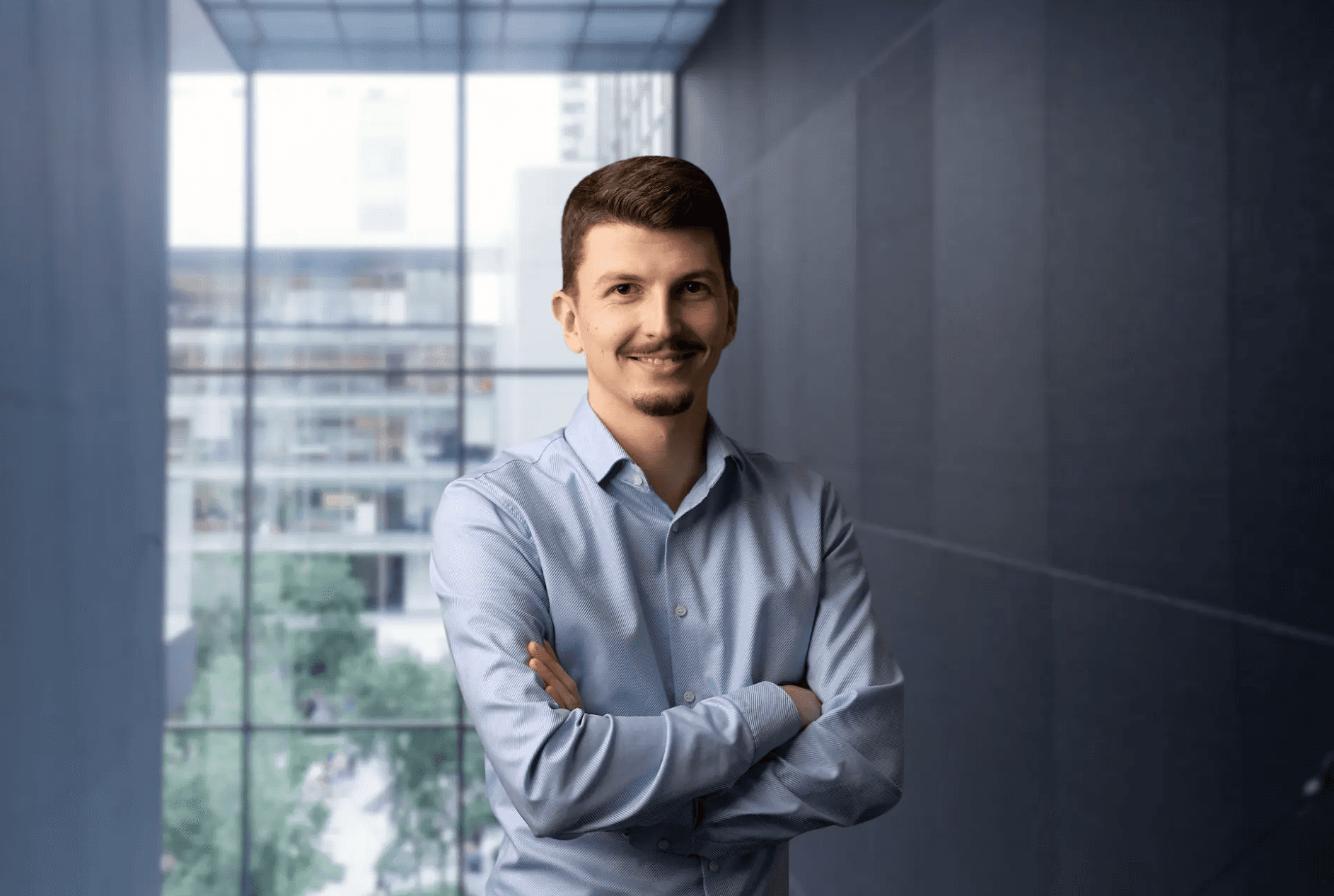 Daniel Antoni, Founder & CEO der VisioSEO GmbH, Portrait eines Mannes mit verschränkten Armen im hellblauen Hemd vor moderner Büroarchitektur mit Glasfassade.