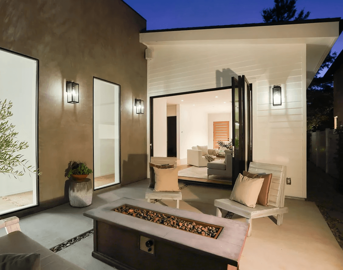 Modern back patio at dusk featuring a rectangular fire pit table and comfortable lounge seating near open glass doors.