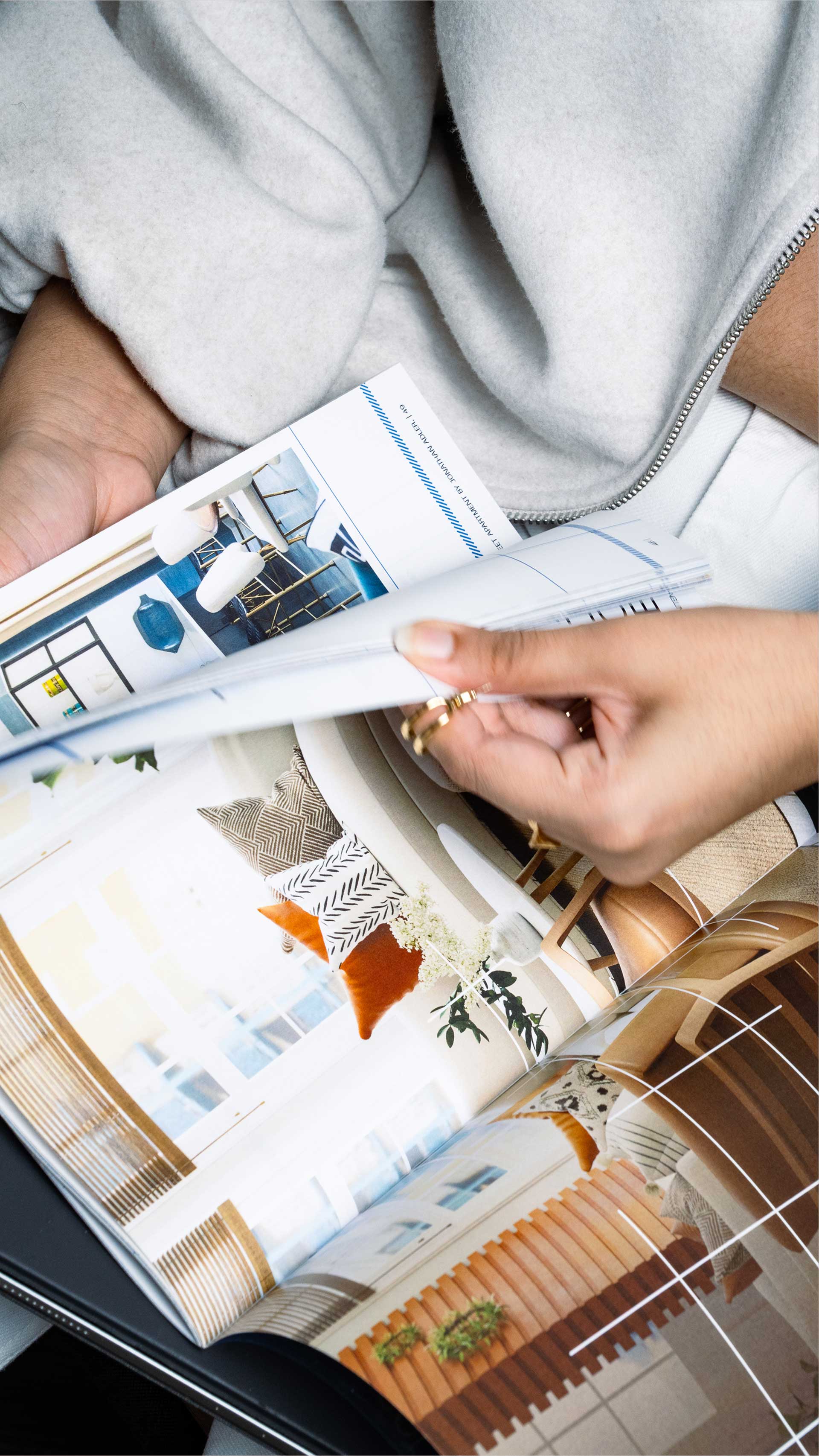 A person wearing a light gray sweater is flipping through a glossy home decor magazine featuring interior design inspirations with images of modern living spaces, including a cozy room accented with orange pillows and contemporary furniture.