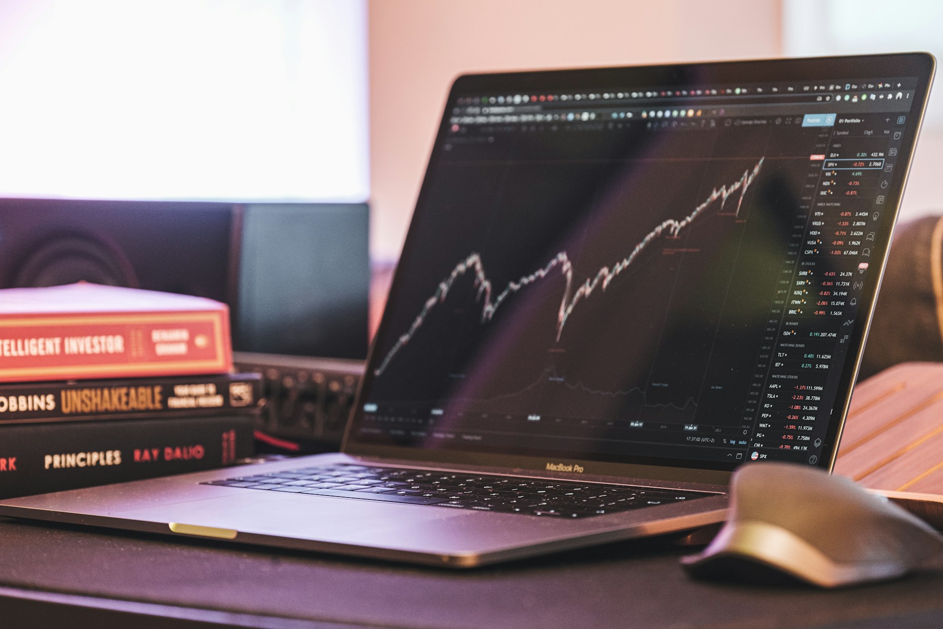 A laptop displaying a stocks trading chart sits on a desk beside books about investing, reflecting a professional trading setup.