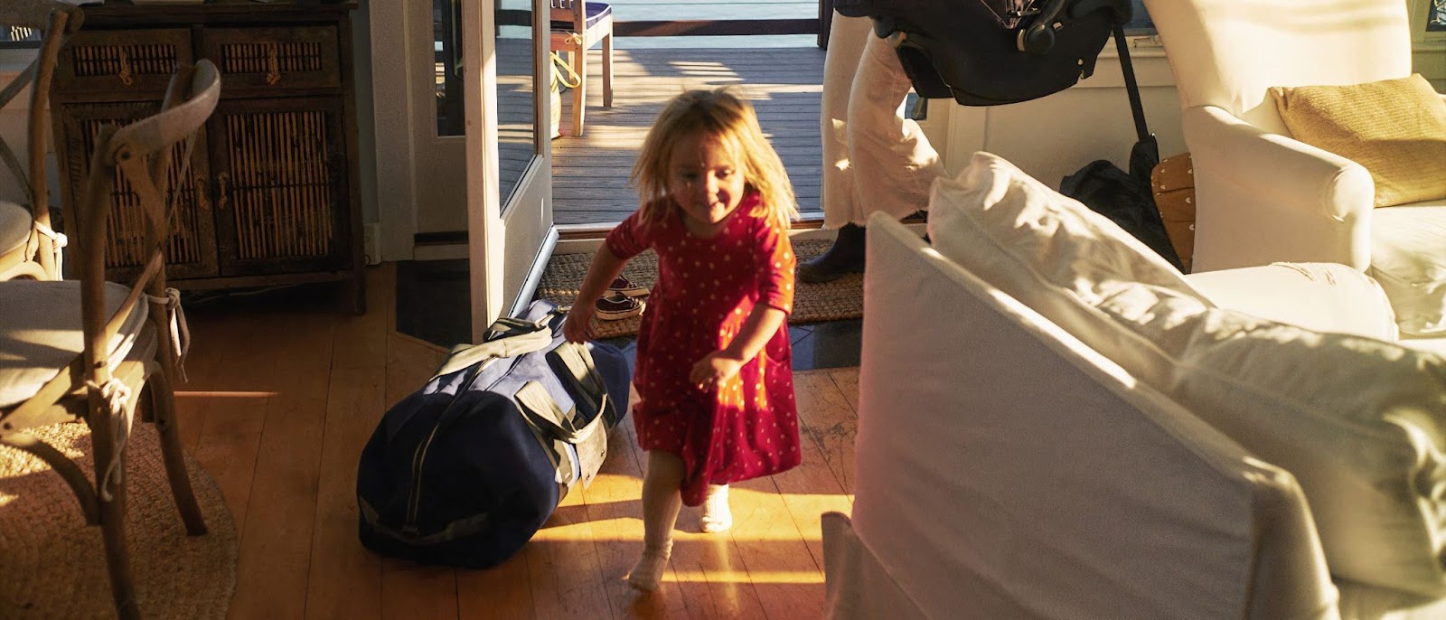 Young girl in red dress runs joyfully inside a sunlit room, near a large blue duffel bag and stroller, with a wooden cabinet and terrace visible.