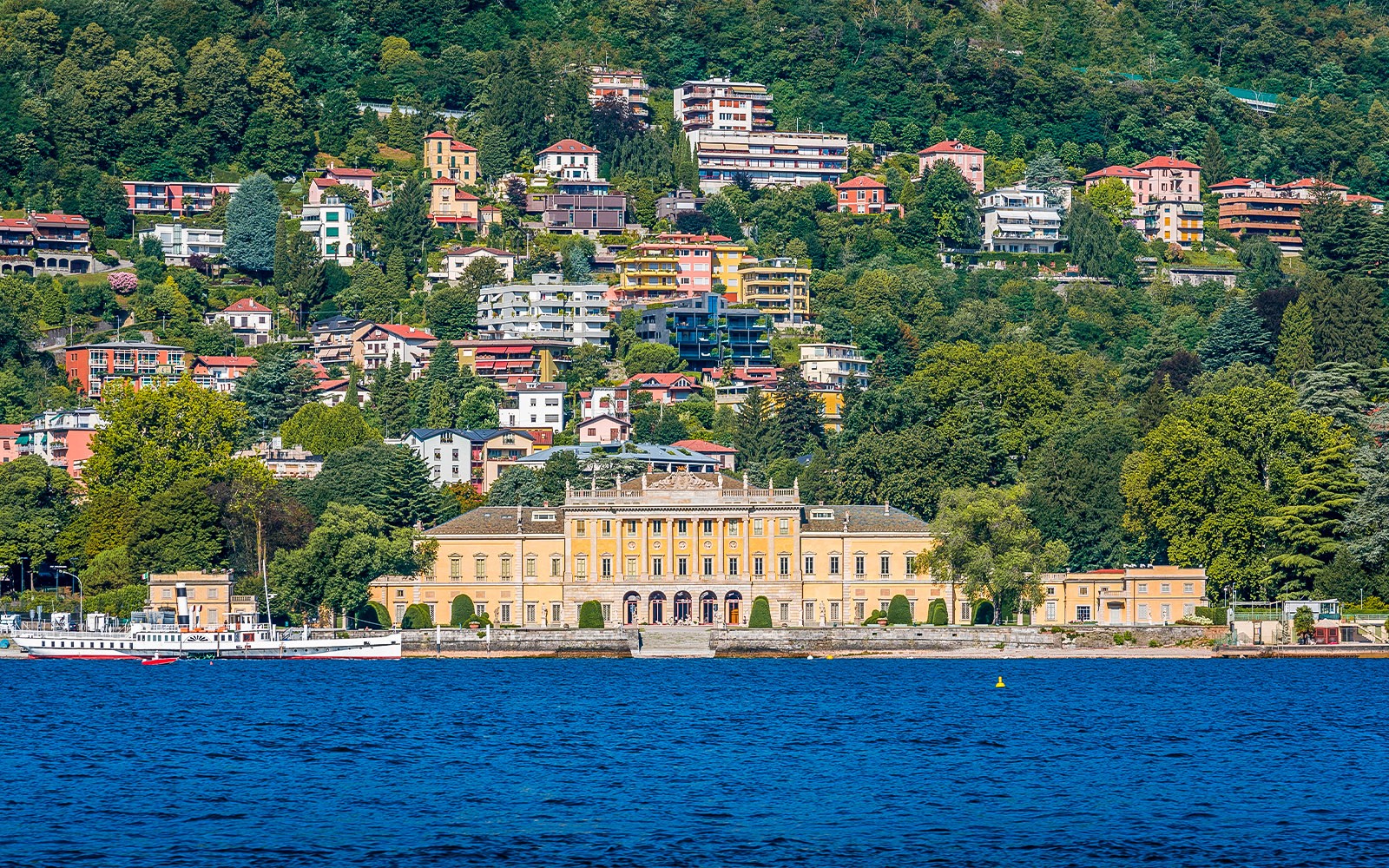 Villa Olmo viewed from a ferry on Lake Como, surrounded by lush hills and colorful buildings, Lombardy, Italy.
