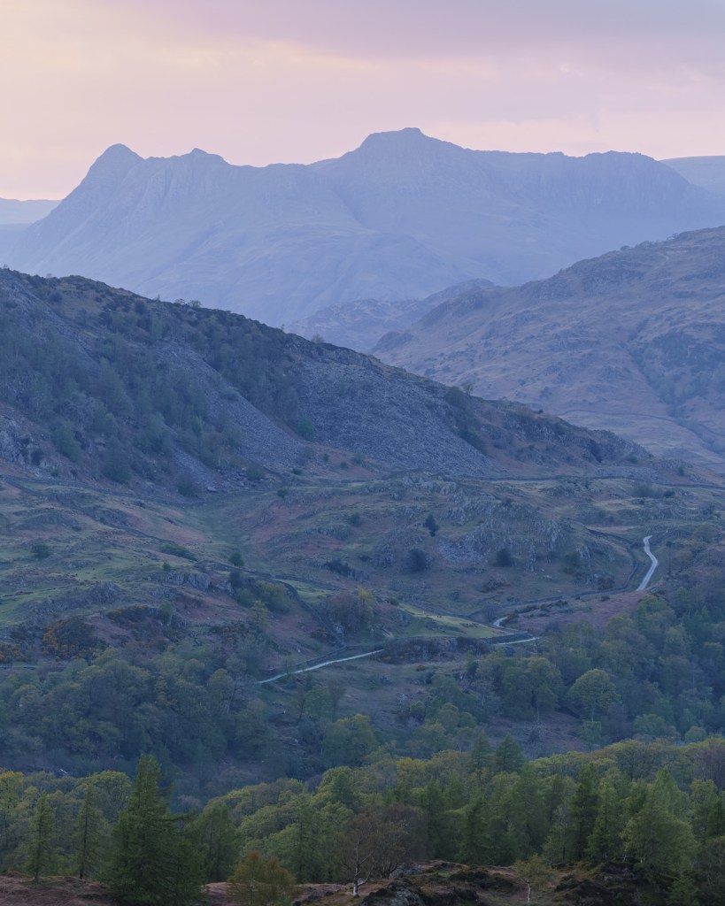 A winding road leading up to Lingmoor. A purple haze covers the distance fells.