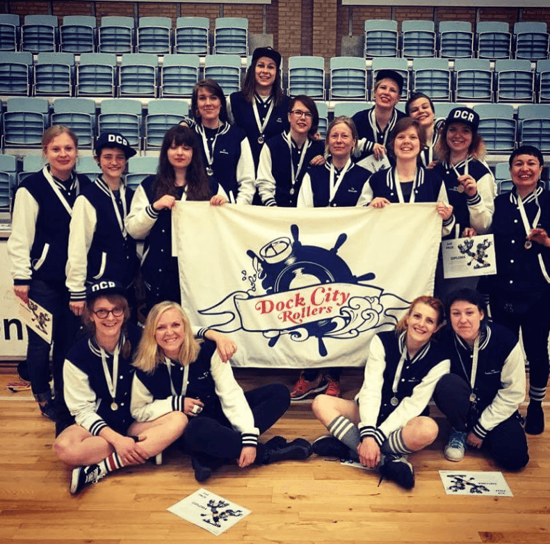 A group of roller derby players posing in front of the camera, holding a flag with the name Dock City Rollers.