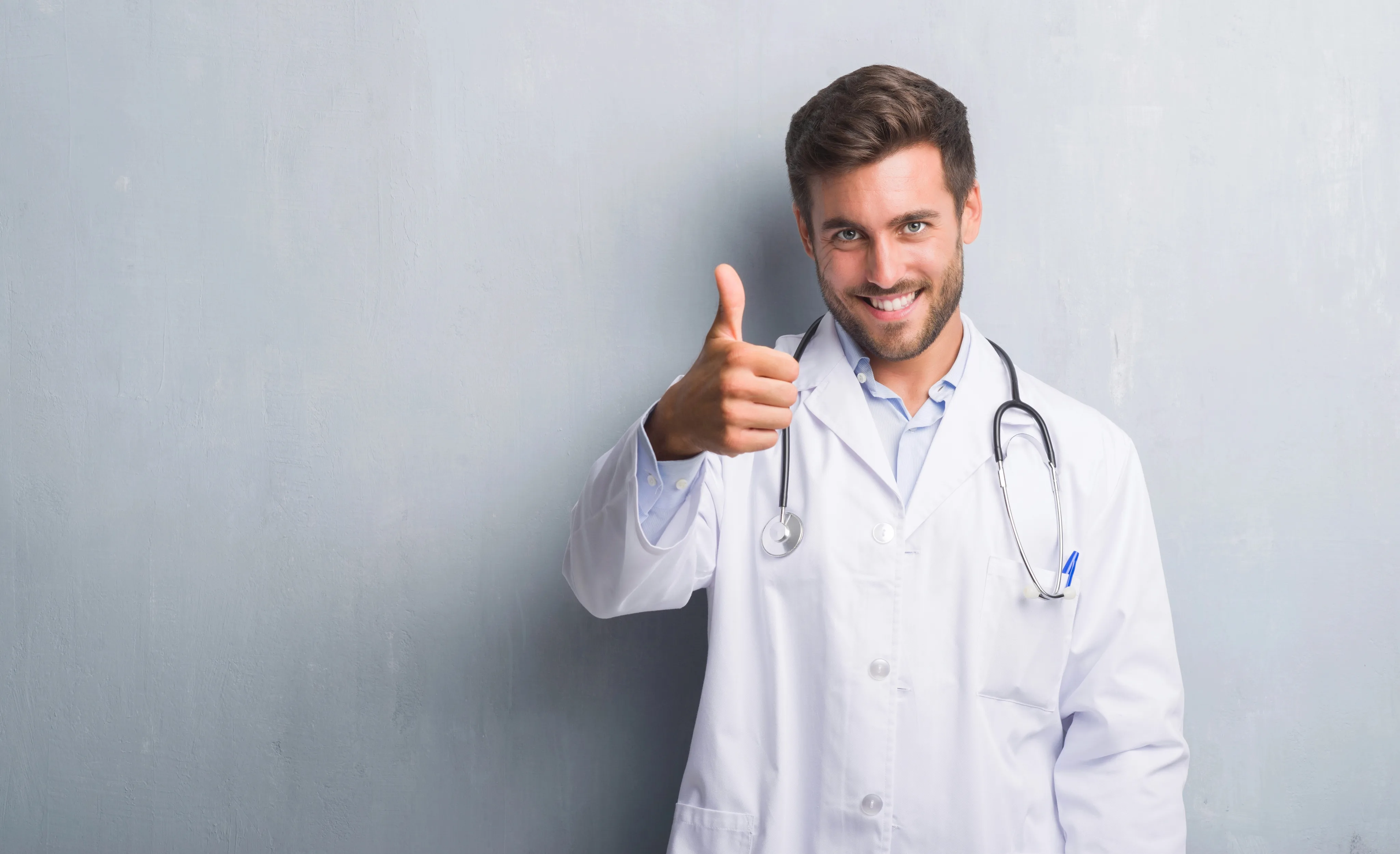 A smiling male doctor in a white lab coat with a stethoscope gives a thumbs-up gesture against a gray background.
