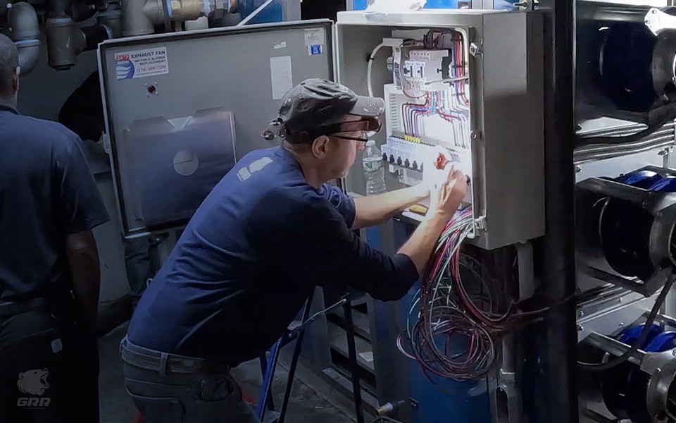 GRR Cooling Experts technician wiring and commissioning a control panel for an EC fan array during a New York HVAC retrofit. The image shows detailed electrical work, open cabinet wiring, and EC fan modules operating in the background inside a mechanical room.